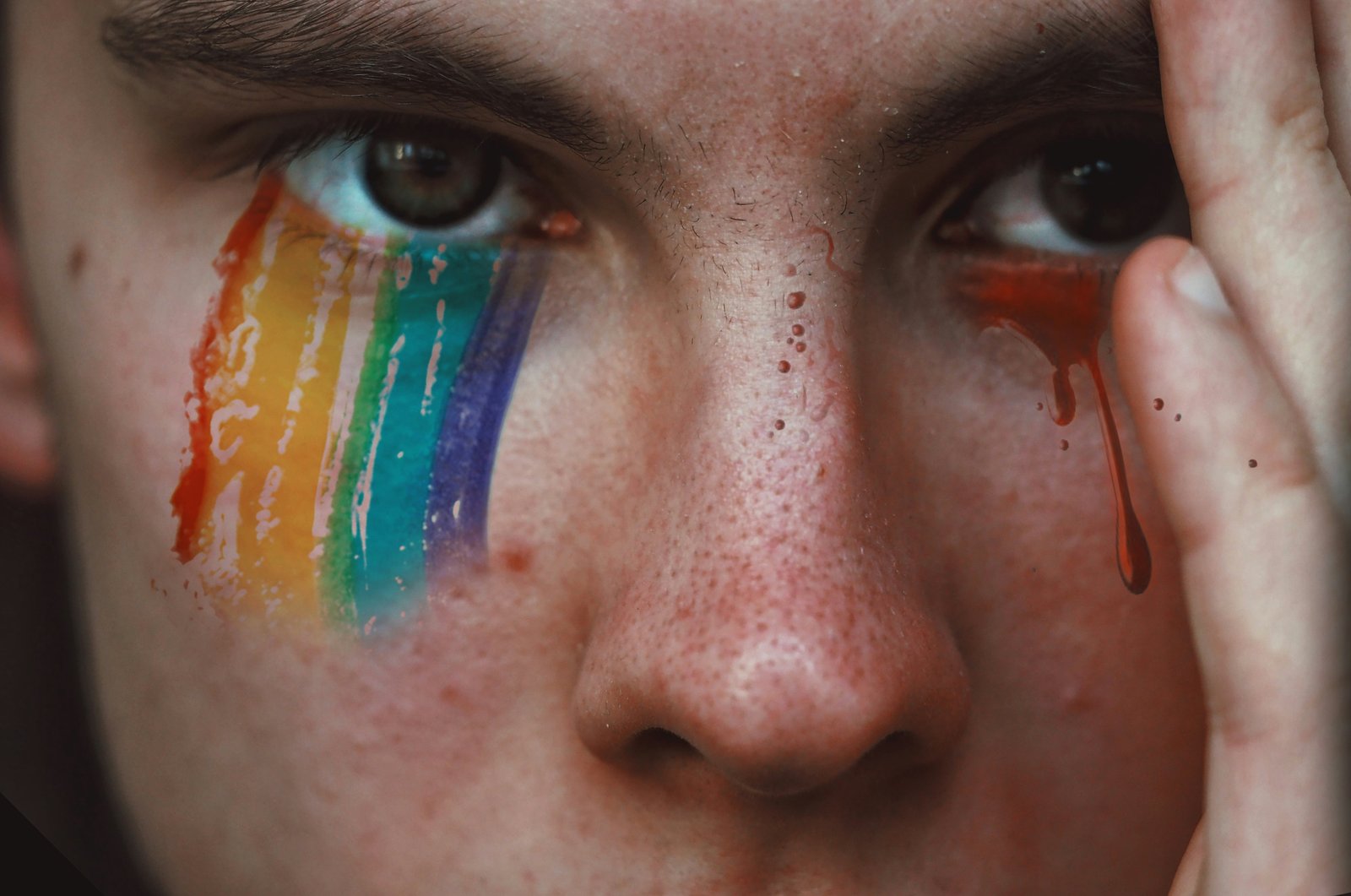 A close-up of a face, one cheek painted with a rainbow, with tears falling